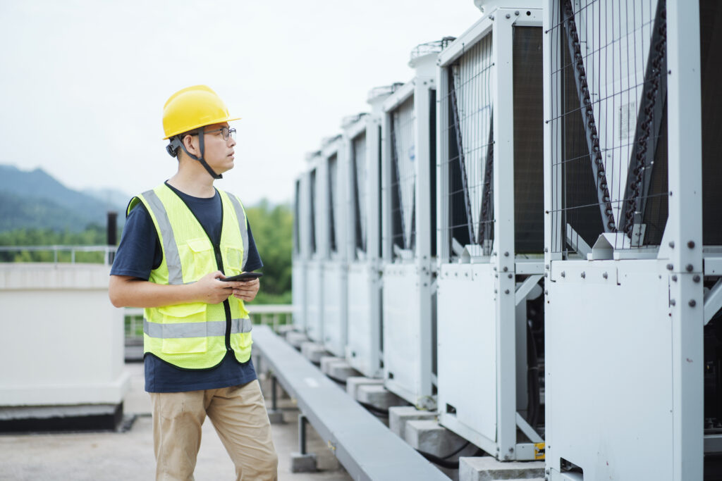 asian engineer working on smart phone with external unit of commercial air conditioning and ventilation system installed on industrial building roof