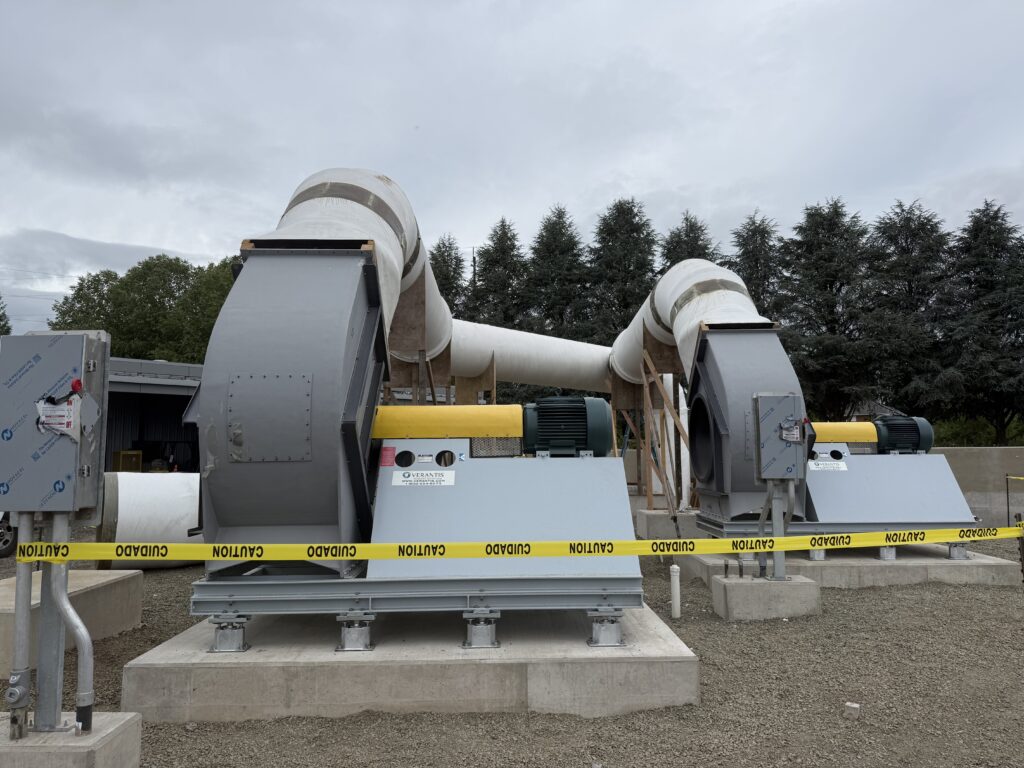 Industrial exhaust fan with white fiberglass duct transition mounted on a concrete pad behind safety barrier tape